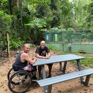 man in wheelchair sitting with a man at a picnic table outdoors in a lush green tropical environment smiling at the camera