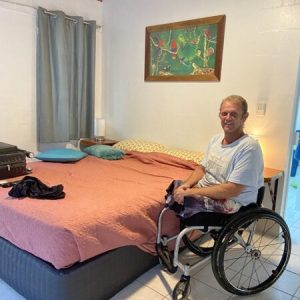 man in wheelchair smiling at the camera next to a made bed in a spacious motel room